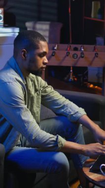 Dikey Video Afrikalı Amerikalı adam oturma odasında makaleler okuyarak ve bilgi toplayarak yeni bir proje için internet kaynaklarını kullanarak araştırma yapıyor. Çoklu görev. Kamera A.