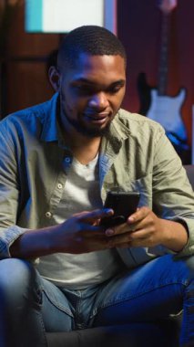 Vertical Video Chill 'in serbest çalışan adamı cep telefonuyla koltukta eğleniyor, arkadaşlarına sosyal medyada mesaj atıyor ve gülüyor. Konforlu oturma odaları uzak yaşam tarzında iş ve eğlence sunar.