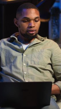 Dikey video odaklı siyahi kişi, şık çatı katında dizüstü bilgisayarda birden fazla iş yapar, internete bağlanır ve günlük serbest işleri ve uzak sorumlulukları yönetir. Kanepede ağ taraması