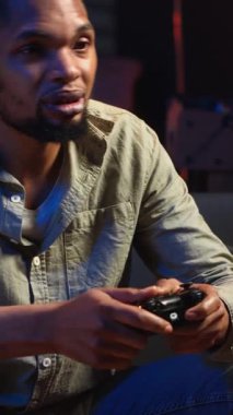 Dikey Video Afrikalı Amerikalı adam boş zamanlarını patlamış mısırla oyun oynayarak geçiriyor, oyun gecesinde evinde eğleniyor. Genç yetişkin, konsolunu televizyona bağlıyor. Kamera.