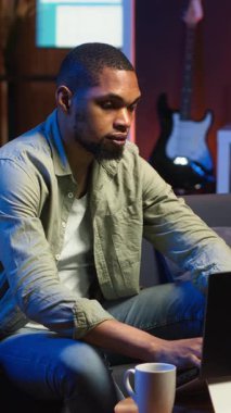 Dikey Video Afrikalı Amerikalı adam oturma odasında makaleler okuyarak ve bilgi toplayarak yeni bir proje için internet kaynaklarını kullanarak araştırma yapıyor. Çoklu görev. Kamera B.