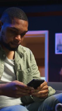 Dikey Görüntü Rahatlatıcı siyah adam rahat bir yerde akıllı telefonuyla kahve masasında oturuyor. Modern ev, şık bir dairede boş zamanların ve çevrimiçi bağlantının altını çiziyor. Kamera.