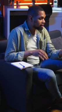 Dikey Video Afrikalı Amerikalı tele-işçi video konferansında iş arkadaşını arayıp not alıyor, yeni fikirler yazıyor ve oturma odasında internet üzerinden bilgi alışverişinde bulunuyor. Ders kitabı verileri. Kamera A.