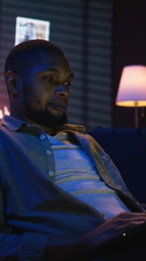 Dikey Video Yetenekli Afro-Amerikan adam havalı oturma odasında dizüstü bilgisayarda çoklu görev yapıyor, serbest çalışma ve uzak sorumlulukları yönetirken internete bağlanıyor. Fasulye torbasında ağ taraması