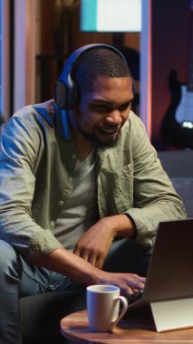 Dikey video odaklı siyah adam oturma odasında dizüstü bilgisayarla telgraf çekerek görevini tamamlamaktan ve başarıya ulaşmaktan memnun. Esnek bir çalışma alanında konfor ve iş verimliliğini dengeleyen uzaktan çalışan