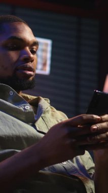 Dikey Video Genç Siyah Adam sosyal medya mesajlaşma uygulamaları üzerinden arkadaşlarına mesaj atıyor, akıllı telefonunu kullanarak internete kayıyor. Odaklanmış biri telefonuyla rahatlıyor, oturma odasında oturuyor.