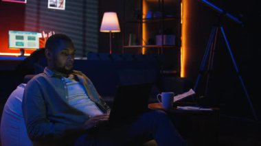 Yetişkin bir adam evdeki fasulye torbasının üzerine yatarken bilgisayarında daktilo yazıyor. Serbest ve çevrimiçi işler yapıyor. Siyahi adam birden fazla iş yapıyor ve uzaktan kumandanın esnekliğini kucaklıyor. Kamera A.