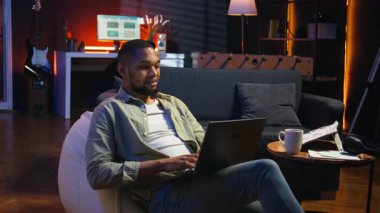 Yetişkin bir adam dizüstü bilgisayarında yazıyor şık çatı katında koltukta yatıyor, serbest ve çevrimiçi işlerle uğraşıyor. Siyahi kişi, uzaktan kumandanın esnekliğini benimsiyor ve birden fazla iş yapıyor. Kamera A.