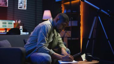 Siyah serbest yazar ödevlerini çözmek, araştırma yapmak ve makaleleri çevrimiçi okumak için ders kitabı üzerine notlar alıyor. Uzaktan kumandalı işten, rahat programdan ve telgraftan hoşlanan genç yetişkinler. Kamera A.