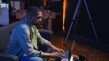 Genç siyahi yetişkin oturma odasında kendi başına çalışıyor, iş programını ayarlıyor ve rahat evinde çalışmaktan keyif alıyor. İnternette dizüstü bilgisayarla gezinen, birden fazla iş yapan biri. Kamera A.