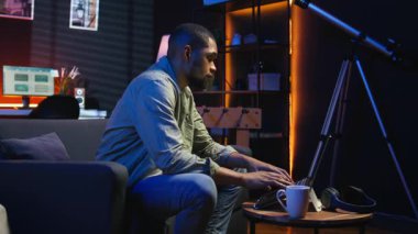 Uzaktan kumandalı bir laptop 'da çalışan Afro-Amerikalı genç bir yetişkin neon ışıkların altında, rahat dairesinde bir kanepede oturuyor. Şık bir evde rahat bir insan modern üretkenliği ön plana çıkarıyor. Kamera A.