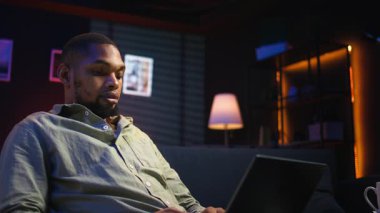 Şık çatı katında dizüstü bilgisayara odaklanmış, internete bağlı, günlük serbest işleri ve uzak sorumlulukları yöneten siyahi kişi. Kanepede ağ taraması. Kamera A.