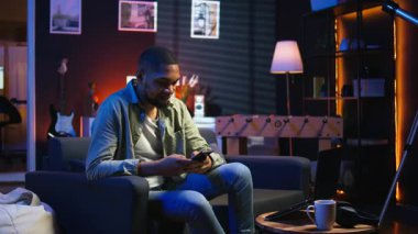 Gülümseyen genç yetişkin arkadaşlarıyla cep telefonunda mesajlaşarak sohbet ediyor, kanepede oturuyor ve sosyal medya uygulamalarını kaydırıyor. Genç adam uzaktan çalışırken eğlenmeye devam ediyor. Kamera A.