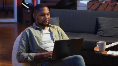 Yetişkin bir adam dizüstü bilgisayarında yazıyor şık çatı katında koltukta yatıyor, serbest ve çevrimiçi işlerle uğraşıyor. Siyahi kişi, uzaktan kumandanın esnekliğini benimsiyor ve birden fazla iş yapıyor. Kamera B.