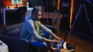 Afrikalı Amerikalı adam oturma odasında makaleler okuyarak ve bilgi toplayarak araştırma yapıyor, yeni bir proje için online kaynakları kullanarak bilgi topluyor. Çoklu görev. Kamera A.