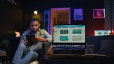 Siyah uzaktan kumandalı işçi ekrandaki analitik ekranın yanında geziniyor, evde oturuyor ve birden fazla cihazla günlük görevleri üzerinde çalışıyor. Çatı katında boş zaman ve iş verimliliği. Kamera A.