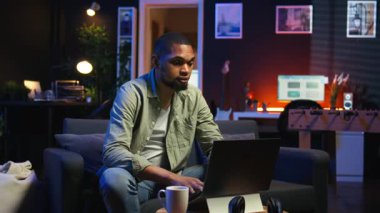 Genç siyahi adam oturma odasında serbest olarak çalışıyor, iş programını ayarlıyor ve rahat evinde çalışmaktan keyif alıyor. İnternette dizüstü bilgisayarla gezinen, birden fazla iş yapan biri. Kamera A.