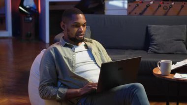 Genç siyahi biri dizüstü bilgisayarında uzaktan kumandalı bir video ile evde oturuyor, görevleri çözmek için online bir konferans web sitesine katılıyor. Oturma odasında telekonferans. Kamera B.