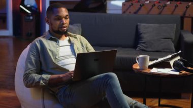 Havalı oturma odasında dizüstü bilgisayarda birden fazla iş yapan yetenekli Afro-Amerikan bir adam, serbest iş ve uzak sorumlulukları yönetirken internete bağlanıyor. Fasulye torbasında ağ taraması. Kamera B.