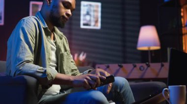 Rahat bir yerde, akıllı telefonuyla kahve masasında oturan rahat siyahi bir adam. Modern ev, şık bir dairede boş zamanların ve çevrimiçi bağlantının altını çiziyor. Kamera B.