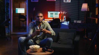 Erkek oyuncu online atış turnuvasındayken patlamış mısır yiyor, evdeki boş zamanlarında video oyunlarıyla eğleniyor. Gevşemiş adam atıştırmalık ve rekabetle oyun gecesinin tadını çıkarıyor. Kamera A.