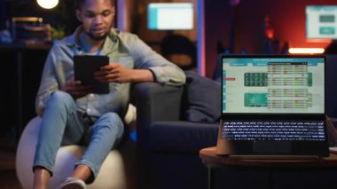 Siyah uzaktan kumandalı işçi ekrandaki analitik ekranın yanında geziniyor, evde oturuyor ve birden fazla cihazla günlük görevleri üzerinde çalışıyor. Çatı katında boş zaman ve iş verimliliği. Kamera B.