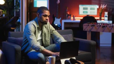 Afrikalı Amerikalı adam oturma odasında makaleler okuyarak ve bilgi toplayarak araştırma yapıyor, yeni bir proje için online kaynakları kullanarak bilgi topluyor. Çoklu görev. Kamera B.