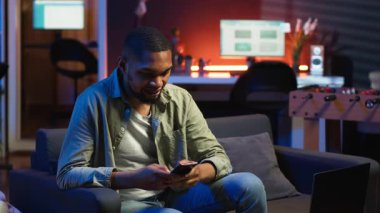 Gülümseyen genç yetişkin arkadaşlarıyla cep telefonunda mesajlaşarak sohbet ediyor, kanepede oturuyor ve sosyal medya uygulamalarını kaydırıyor. Genç adam uzaktan çalışırken eğlenmeye devam ediyor. Kamera B.