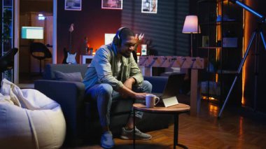 Oturma odasında dizüstü bilgisayarla çalışan odaklanmış siyahi adam görev tamamlanınca mutlu oluyor ve başarıya ulaşıyor. Uzaktan kumandalı işçi, esnek bir çalışma alanında rahatlık ve iş verimliliğini dengeliyor. Kamera A.