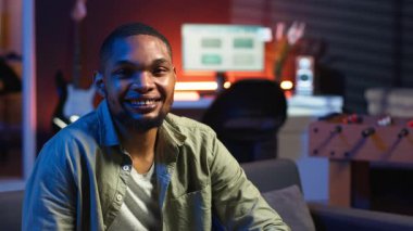 Kanepeye uzanmış, telefonda mesajlaşan, modern dairesinde rahatlayan ve gülümseyen siyahi serbest yazar portresi. Oturma odasından havalı ışıklarla günlük tele-çalışma görevlerini çözen bir adam. Kamera B.