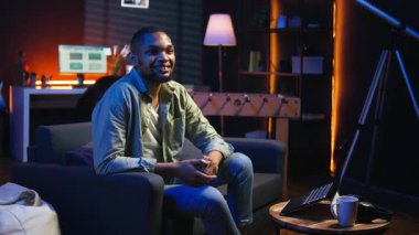 Kanepeye uzanmış, telefonda mesajlaşan, modern dairesinde rahatlayan ve gülümseyen siyahi serbest yazar portresi. Oturma odasından havalı ışıklarla günlük tele-çalışma görevlerini çözen bir adam. Kamera A.