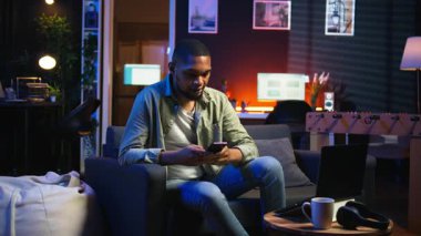 Afro-amerikalı bir insan koltukta akıllı telefon uygulaması üzerinden mesaj atıyor, evde dinlenmeden önce görevlerini tamamlıyor. Genç yetişkin cep telefonunu kaydırıyor, mesajlarla sohbet ediyor. Kamera A.