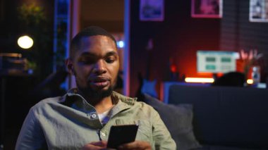 Afro-Amerikalı genç adam telefonunu kanepeye bırakıp, arkadaşlarına sosyal medyada mesaj atıp eğleniyor. Modern oturma odası, uzak yaşam tarzında boş vakitleri gösteriyor. Kamera A.