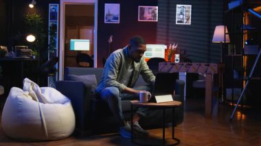 Yetişkin bir adam dizüstü bilgisayarda yazıyor şık bir dairede koltukta yatıyor, serbest çalışıyor ve internetten çalışıyor. Siyahi kişi birden fazla iş yapıyor ve uzaktan kumandanın esnekliğini kucaklıyor. Kamera A.