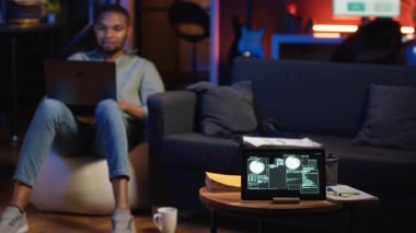 Çatı katında siber güvenlik üzerinde çalışan bir siyahi bilişim uzmanı. Cihazlarla, hackleme girişimleriyle, şifreleme ve siber savunmayla uğraşıyor. Ekranda güvenlik ihlali uyarısı, veri güvenliği riskleri görünüyor. Kamera B.