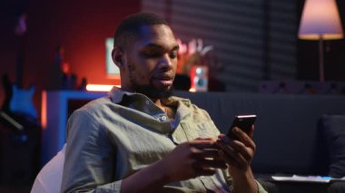 Afro-Amerikalı genç adam telefonunu kanepeye bırakıp, arkadaşlarına sosyal medyada mesaj atıp eğleniyor. Modern oturma odası, uzak yaşam tarzında boş vakitleri gösteriyor. Kamera B.