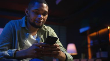 Sıradan bir Afro-Amerikan erkeği rahat bir yerde akıllı telefonuyla kahve masasında oturuyor. Modern ev, lüks bir çatı katında boş vakit ve çevrimiçi bağlantıyı ön plana çıkarıyor. Kamera A.