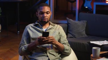 Genç siyahi adam sosyal medya mesajlaşma uygulamaları üzerinden arkadaşlarına mesaj atıyor, akıllı telefonunu kullanarak internete kayıyor. Odaklanmış biri telefonuyla rahatlıyor, oturma odasında oturuyor. Kamera B.