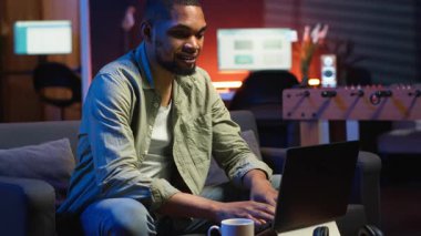 Genç siyahi yetişkin oturma odasında kendi başına çalışıyor, iş programını ayarlıyor ve rahat evinde çalışmaktan keyif alıyor. İnternette dizüstü bilgisayarla gezinen, birden fazla iş yapan biri. Kamera B.