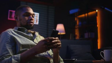 Genç siyahi adam sosyal medya mesajlaşma uygulamaları üzerinden arkadaşlarına mesaj atıyor, akıllı telefonunu kullanarak internete kayıyor. Odaklanmış biri telefonuyla rahatlıyor, oturma odasında oturuyor. Kamera A.