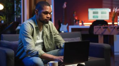 Genç yetişkin kanepede oturuyor ve analitikle araştırma kağıtları okuyor, evdeki dosya ve belgelerle birden fazla iş yapıyor. Adam profesyonel ya da kişisel gelişim konularına dalıyor. Kamera B.