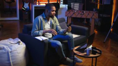 Afrikalı Amerikalı tele-işçi video konferansında iş arkadaşını arayıp notlar alıyor, yeni fikirler yazıyor ve oturma odasında internet görüşmeleri hakkında bilgi alışverişinde bulunuyor. Ders kitabı verileri. Kamera A.