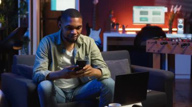 Rahat takılan, cep telefonuyla kanepede dinlenen, sosyal medyada arkadaşlarına mesaj atan ve gülen bir adam. Konforlu oturma odası gösterileri, uzak yaşam tarzında iş ve eğlence. Kamera B.