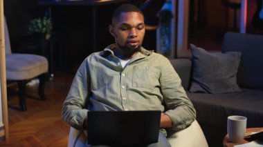 Şık çatı katında dizüstü bilgisayara odaklanmış, internete bağlı, günlük serbest işleri ve uzak sorumlulukları yöneten siyahi kişi. Kanepede ağ taraması. Kamera B.