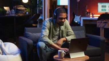 Oturma odasında dizüstü bilgisayarla çalışan odaklanmış siyahi adam görev tamamlanınca mutlu oluyor ve başarıya ulaşıyor. Uzaktan kumandalı işçi, esnek bir çalışma alanında rahatlık ve iş verimliliğini dengeliyor. Kamera B.
