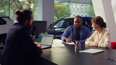 Araba galerisi satıcısı müşterileriyle aynı masada oturuyor. Test sürüşünü bitirdikten sonra imzalamaları için sözleşme imzalıyor. Ajan satın almak için fazladan araç özellikleri öneriyor, imza için bekliyor, kamera B