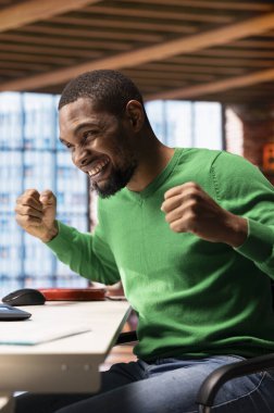 Memlekette başarı, tezahürat ve el kol hareketi ile heyecanlanan çok mutlu bir adam. Mutlu Afro-Amerikalı insan gülümsüyor, başarıyı kutluyor, olumlu duygular gösteriyor, apartman masasında oturuyor.