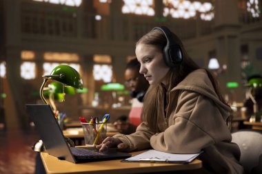 Üniversite öğrencisi, araştırma için mükemmel bir ortam yaratmak için kulaklıkla terapi müziği dinliyor, diploma ödevi üzerinde çalışıyor. Genç kız bilgi toplamak için odak müziği kullanıyor..