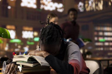 Afrika kökenli Amerikalı bir öğrenci uzun bir gece okuduktan, üniversiteyi bitirmeye ve sınavları geçmeye çalıştıktan sonra kitap yığınının üzerinde uyuyor. Aşırı yüklemeden dolayı kütüphanede dinlenen uykusuz öğrenci..