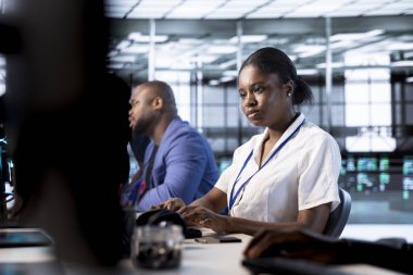 Veri merkezinin elektronik yaşam süresini uzatmak ve devre dışı kalmaktan kaçınmak için yıllık check-up yapan meslektaşlarının yanındaki Afrikalı Amerikalı yönetici. Sunucu merkezindeki kadın sunucu altyapı bileşenlerini izliyor